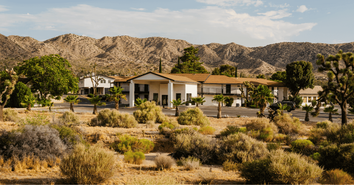 A landscape photo of buildings nestled in a desert area. The buildings are white with tan roofs, and are surrounded by trees and shrubs, with hills or dunes in the background.