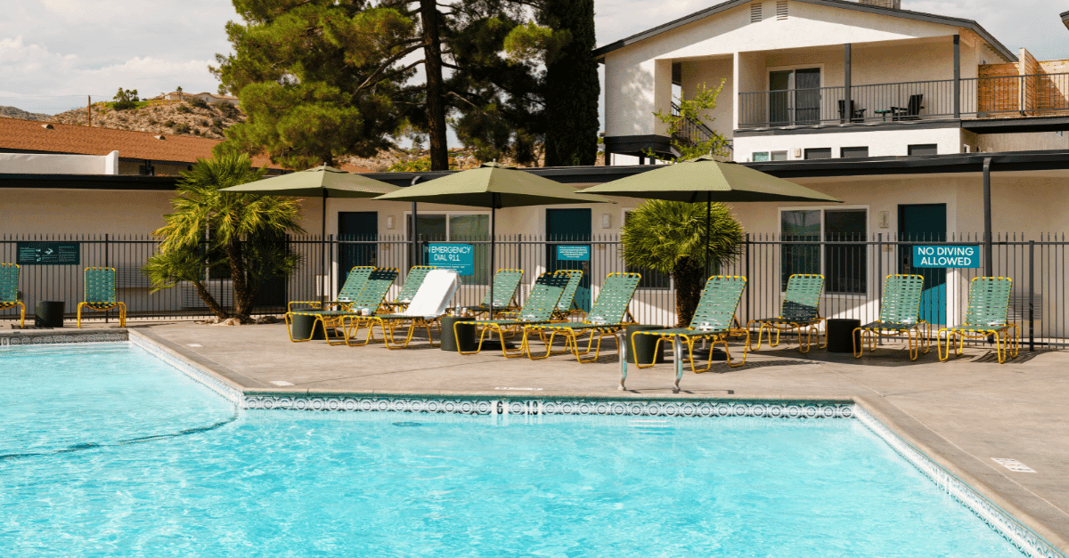 A sunny hotel pool area with a bright, clear pool, and several teal and orange lounge chairs with green square umbrellas in between.