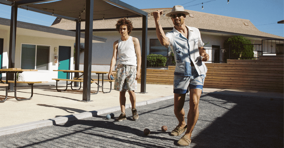 Two men dressed in casual summer shirts and shorts are outside in a sunny space with picnic tables, playing a game with 3 small balls