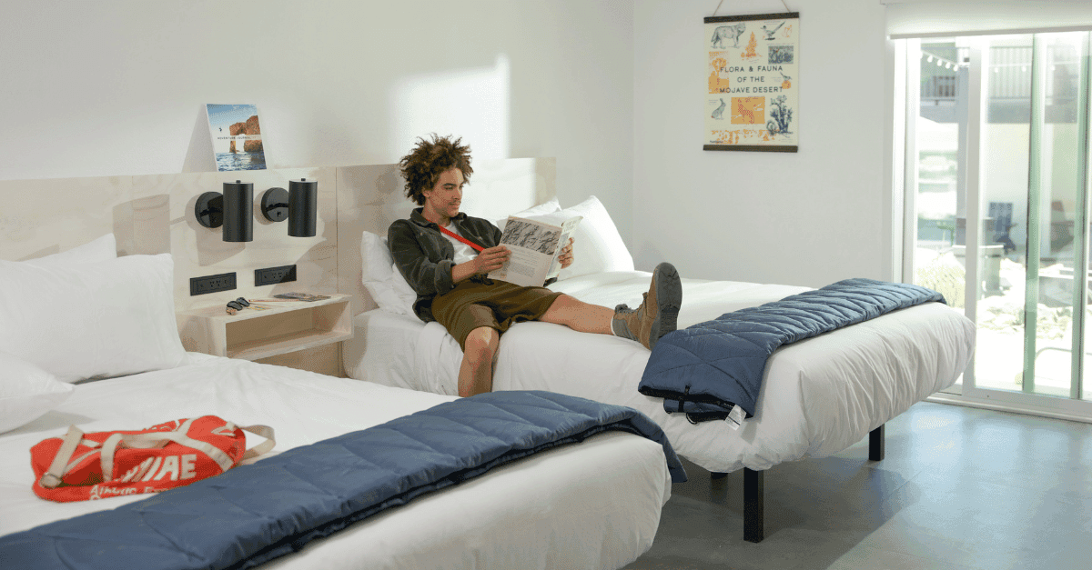 A man lounges on one of two large beds in a white hotel room. He is reading a magazine.