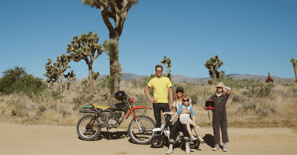 A man, woman, and two blonde little girls are posed in a group with a large dirtbike and a small dirtbike in the desert, there are trees in the background.