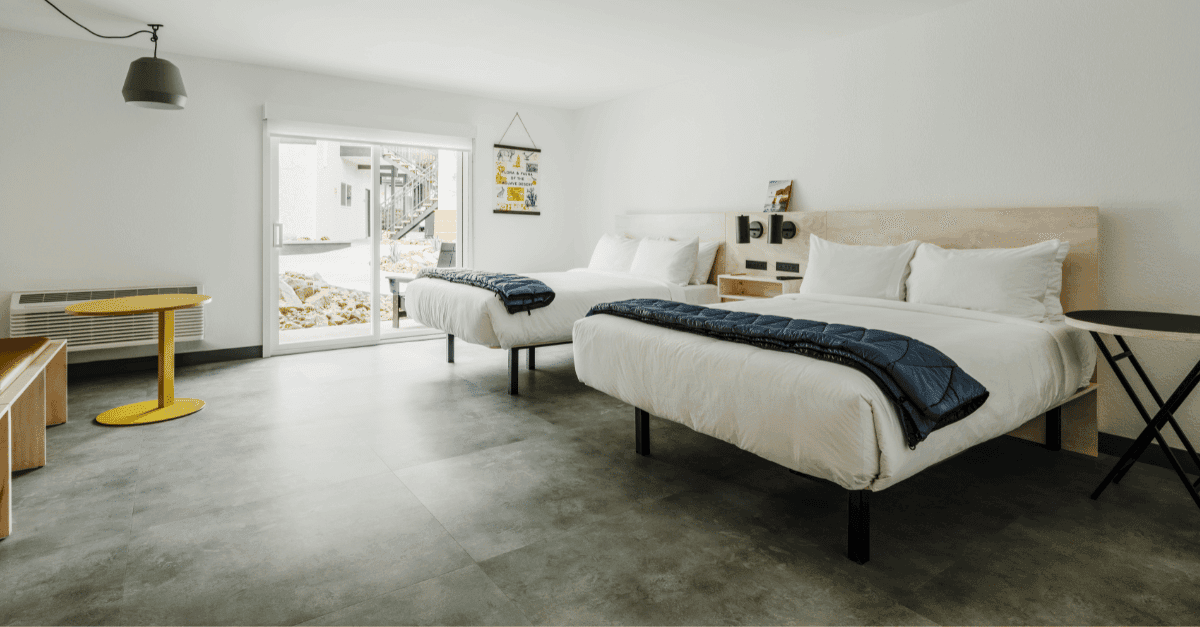 A hotel room with white walls, grey floors, and a patio door. There are 2 large beds next to each other.