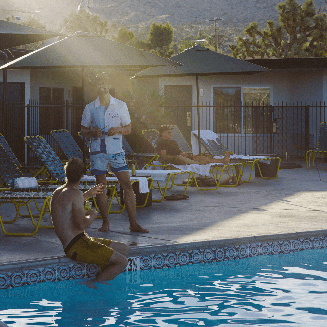 A smiling man is standing and having a conversation by the pool with another man who is seated with his feet in the water. A third man is on a lounge chair in the background.