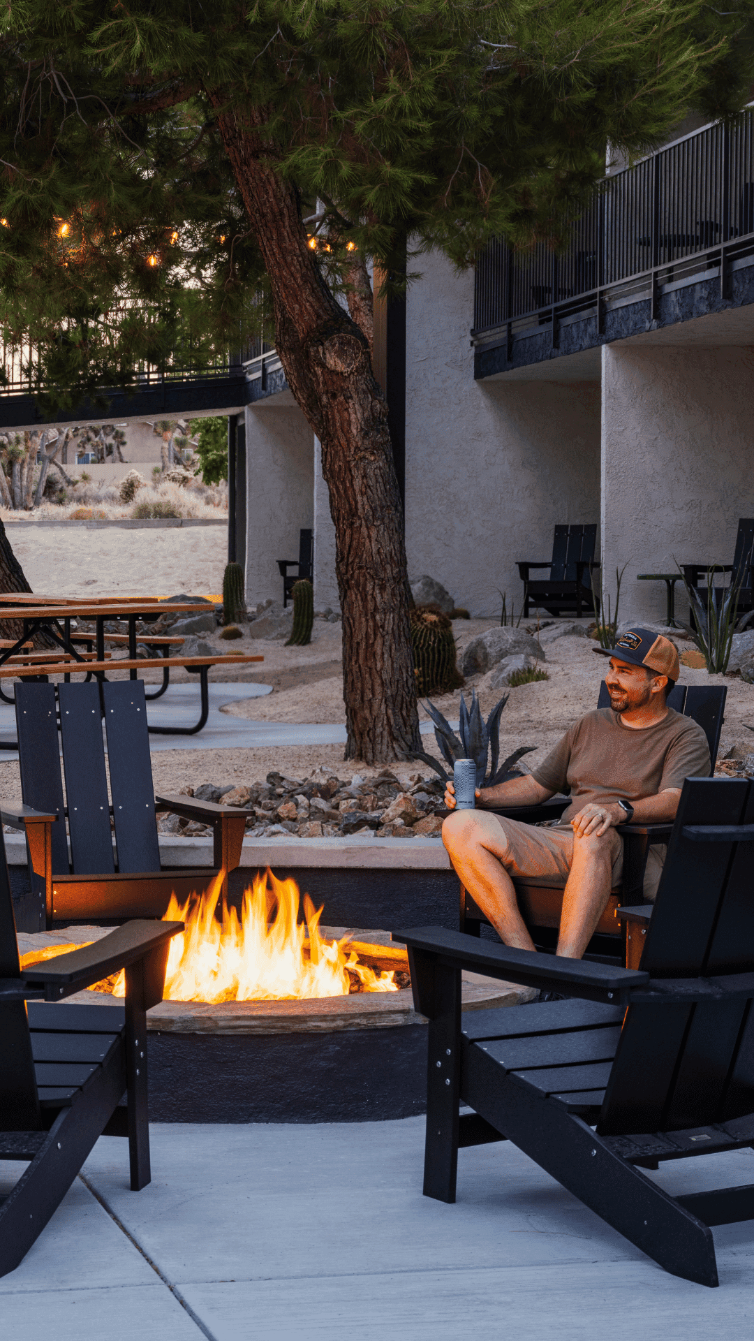 A smiling man in shorts, a tshirt and ball cap sits on a black chair in front of a fire pit. There are 3 other empty black chairs, and a large tree in the background.