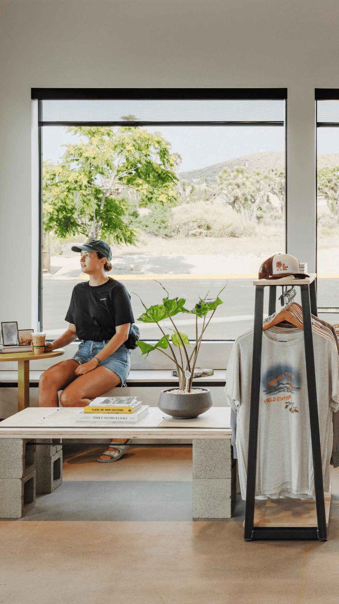 A person in a black shirt, shorts and ball cap sits on a bench in a clean white cafe with a large window, and is holding an iced coffee. A rack of tshirts is nearby.