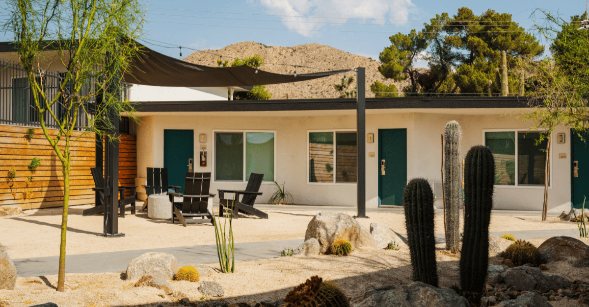 A white hotel building with cacti in the foreground, as well as firepit with chairs in front of the building.