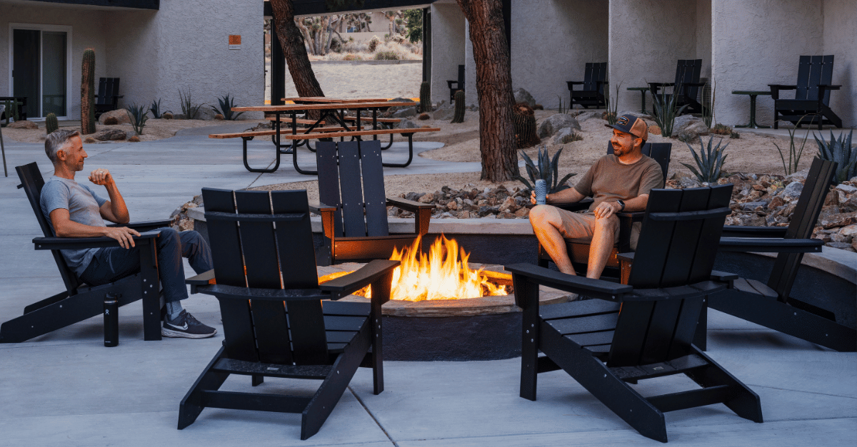 Two men in shorts and t-shirts are sitting in black wooden chairs around a fire pit. There are 3 other empty chairs, with additional chairs and tables in the background.