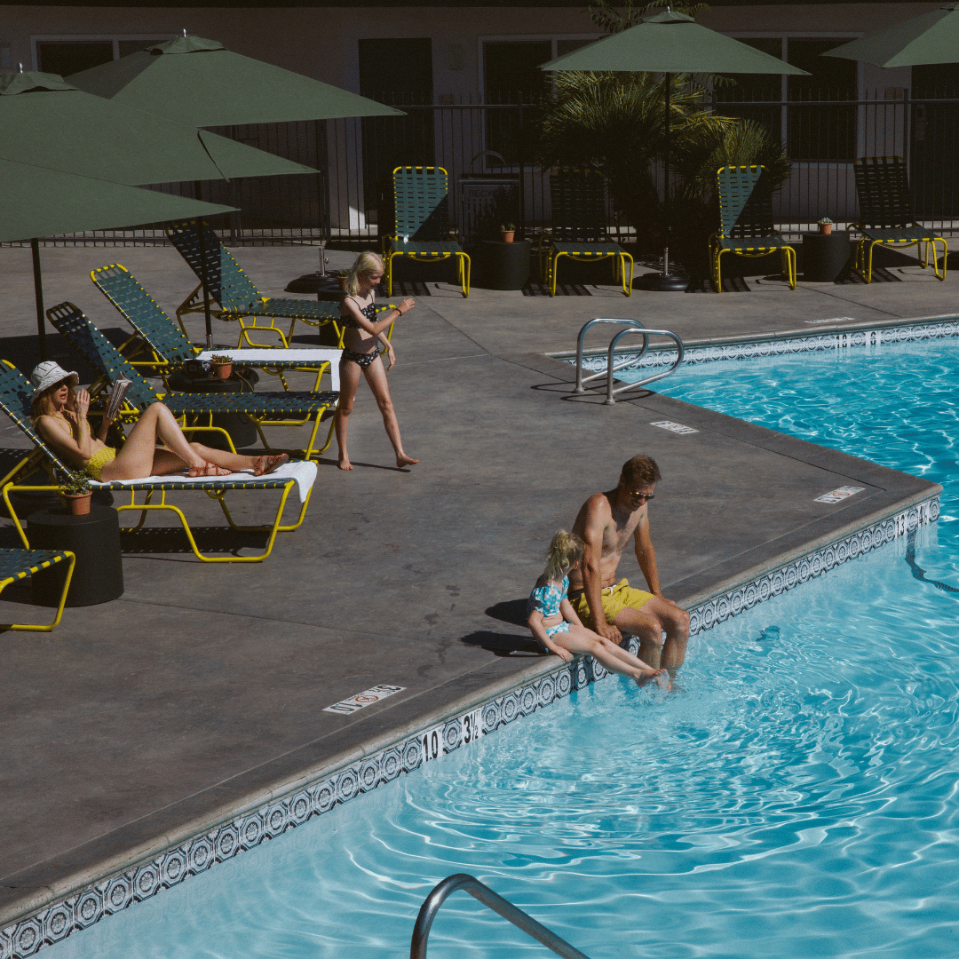 A man and girl sit with their feet in a pool. There are lounge chairs around the pool and one has a woman on it, reading.