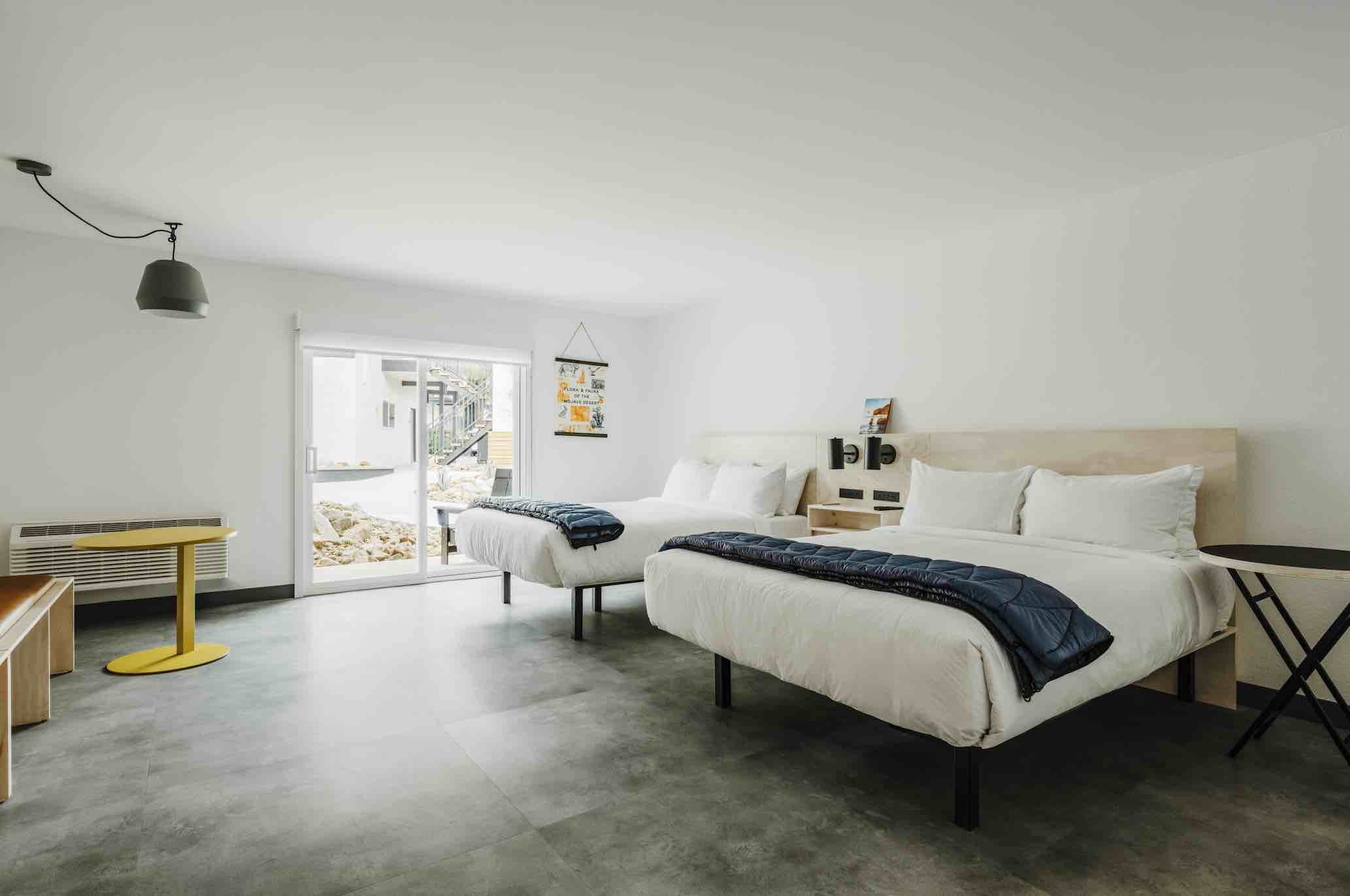 A hotel room with white walls, grey floors, and a patio door. There are 2 large beds next to each other.