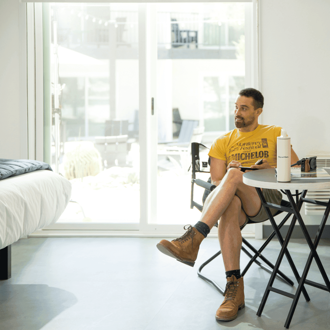 A man sits at a small table in front of a patio door in a white hotel room.