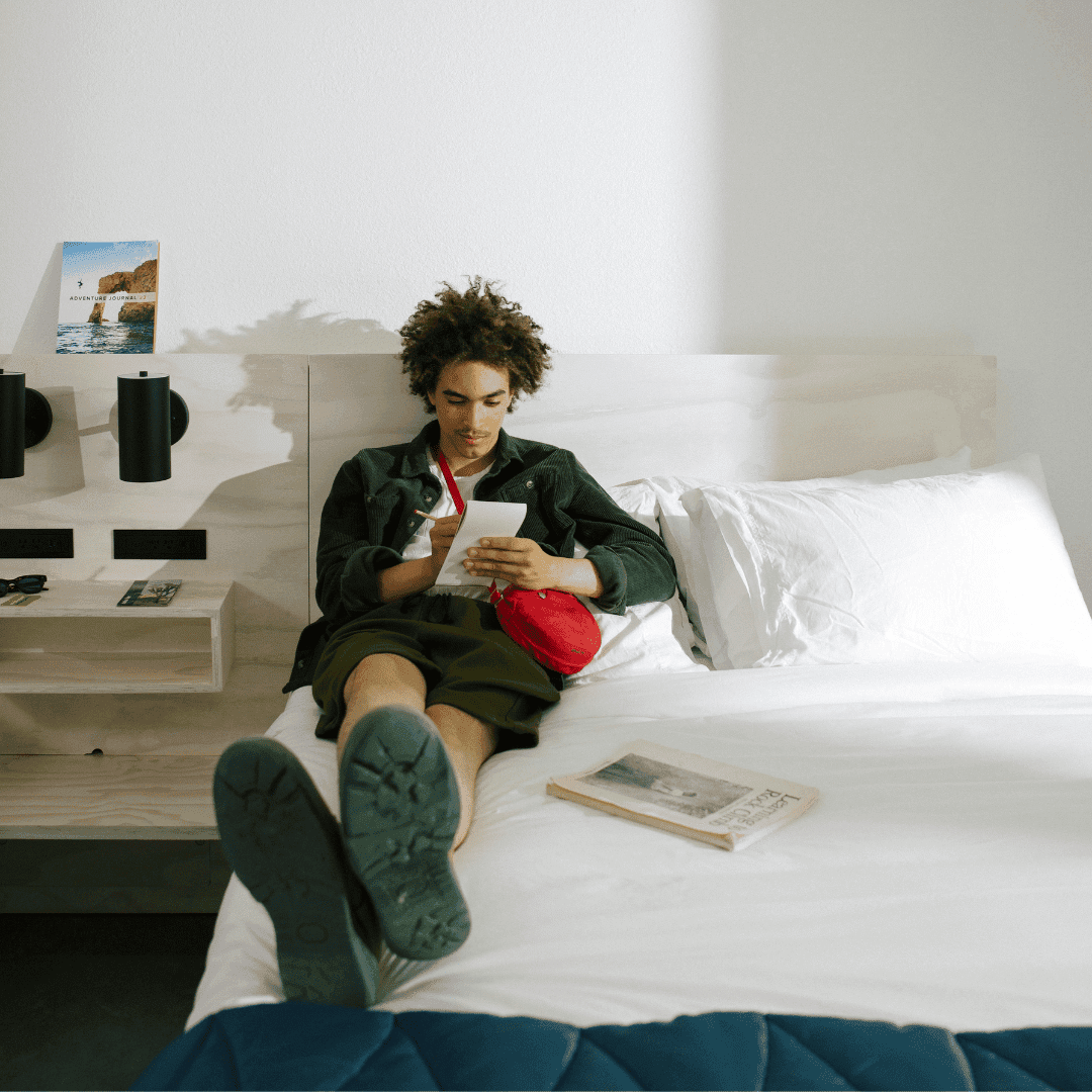 A man lounges on a large bed in a white hotel room. He is writing in a notebook.