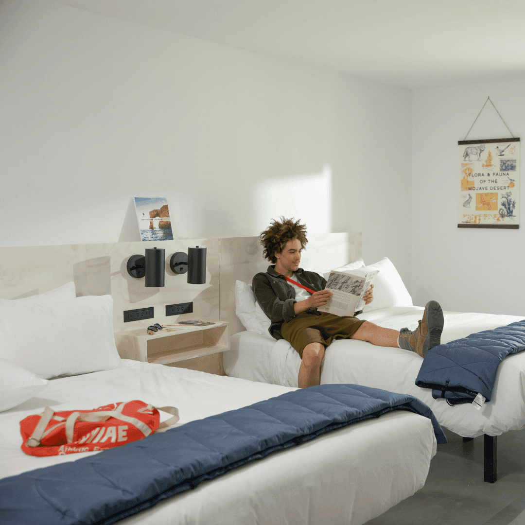 A man lounges on one of two large beds in a white hotel room. He is reading a magazine.