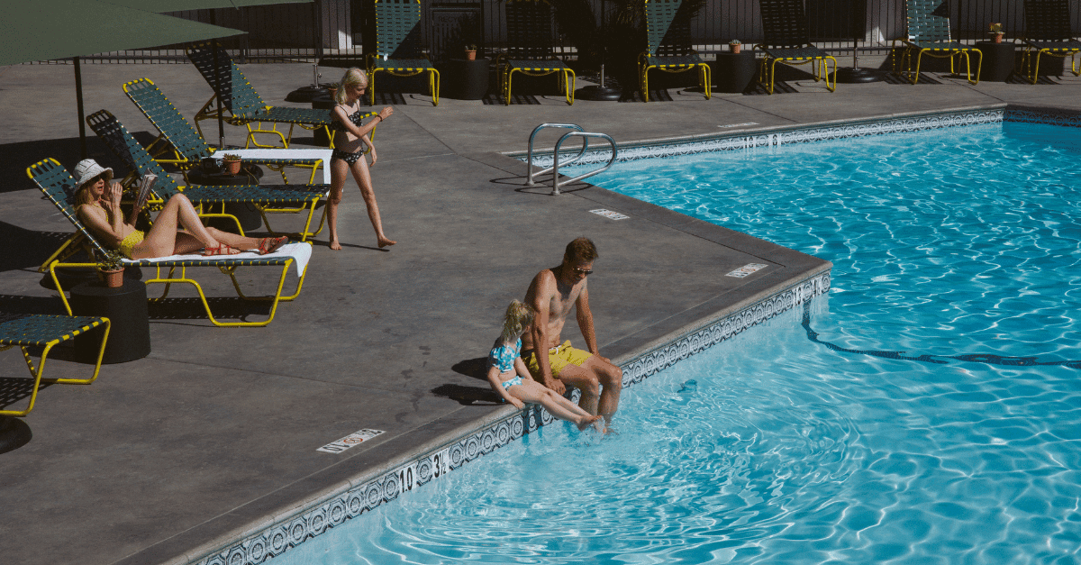 A man and girl sit with their feet in a pool. There are lounge chairs around the pool and one has a woman on it, reading.