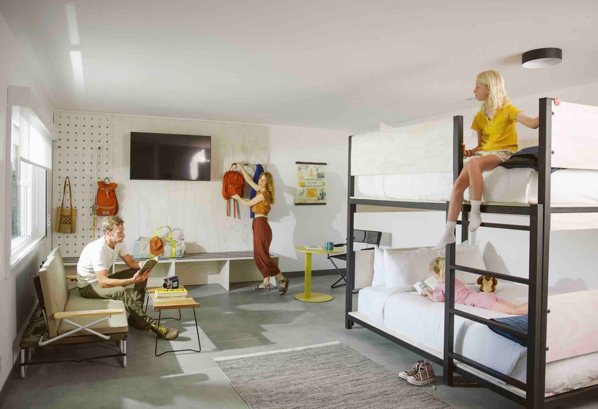 A man, woman, and 2 little blonde girls are hanging out in a room with twin beds, a TV, a couch, and tables. The man is reading and the girls are on the two beds, while the woman is picking up a backpack off a hook.