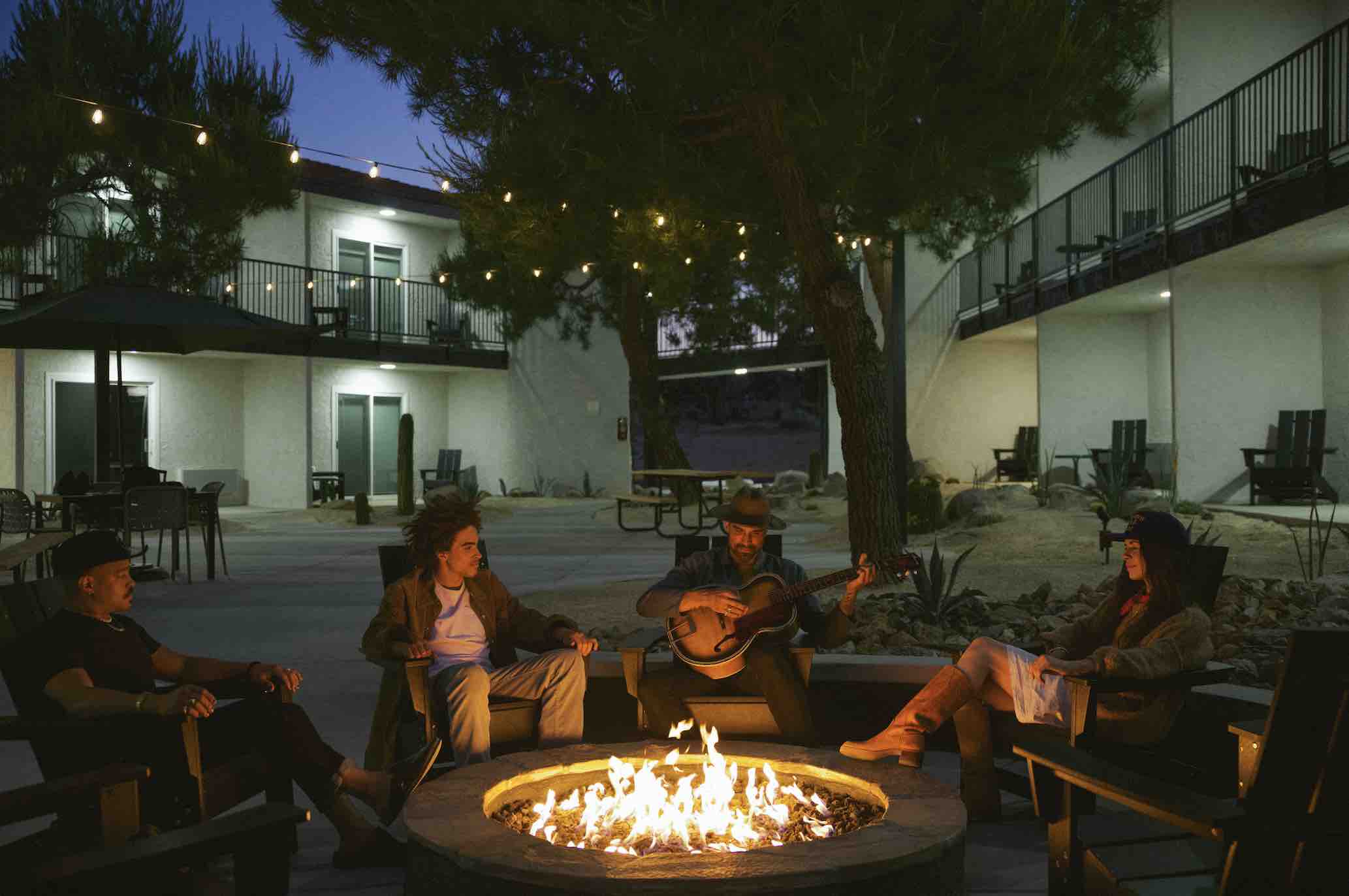 Four people sitting around a fire pit at night, the glow from the fire lights them. A man in a hat is playing guitar.