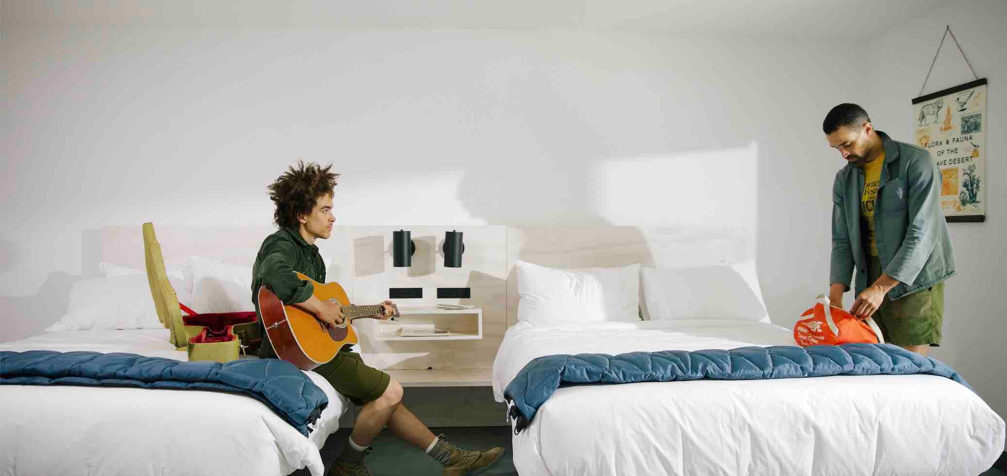 Two men are in a white hotel room with two large beds. One is sitting on a bed playing the guitar, the other is packing a bag.