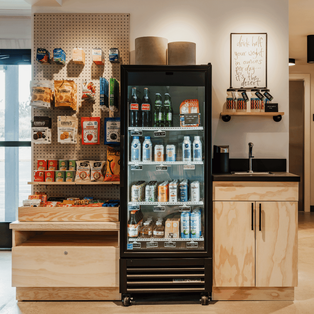 A merchandise area with snacks and drinks. The snacks are on a ooden shelf and the drinks are in a cooler case. There is also a small sink with cupboard underneath.
