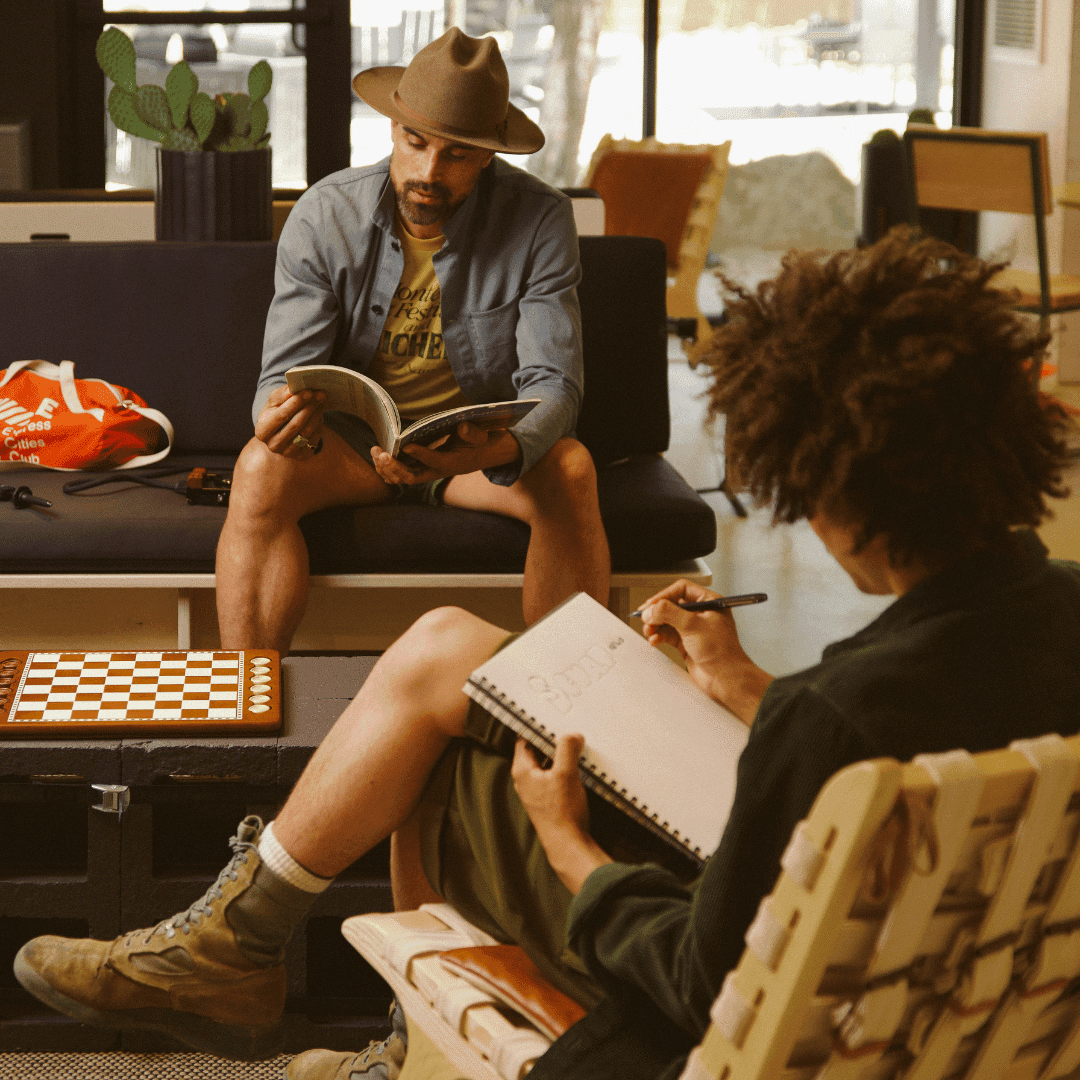 Two people sitting on couches in a cafe. One is reading and one is writing in a notebook.