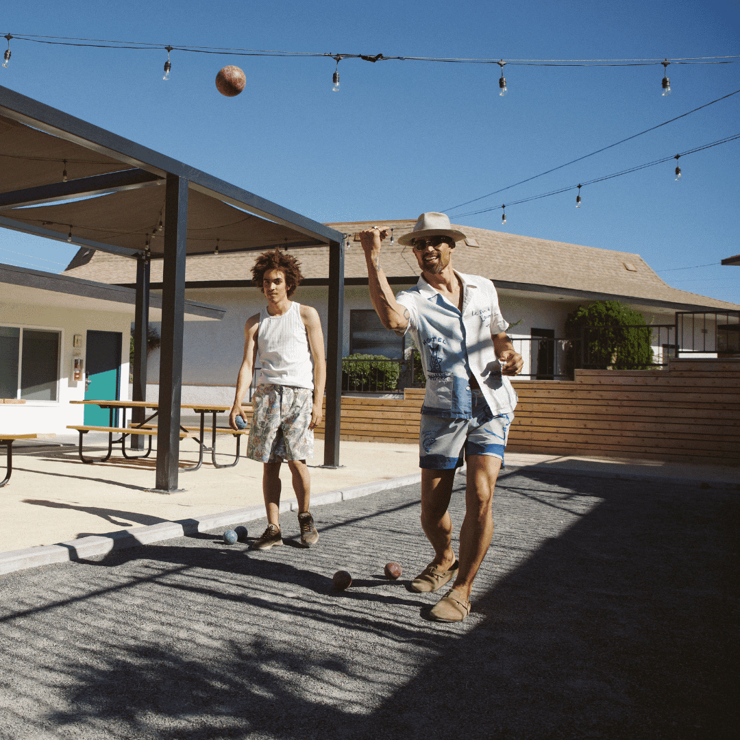Two men dressed in casual summer shirts and shorts are outside in a sunny space with picnic tables, playing a game with 3 small balls