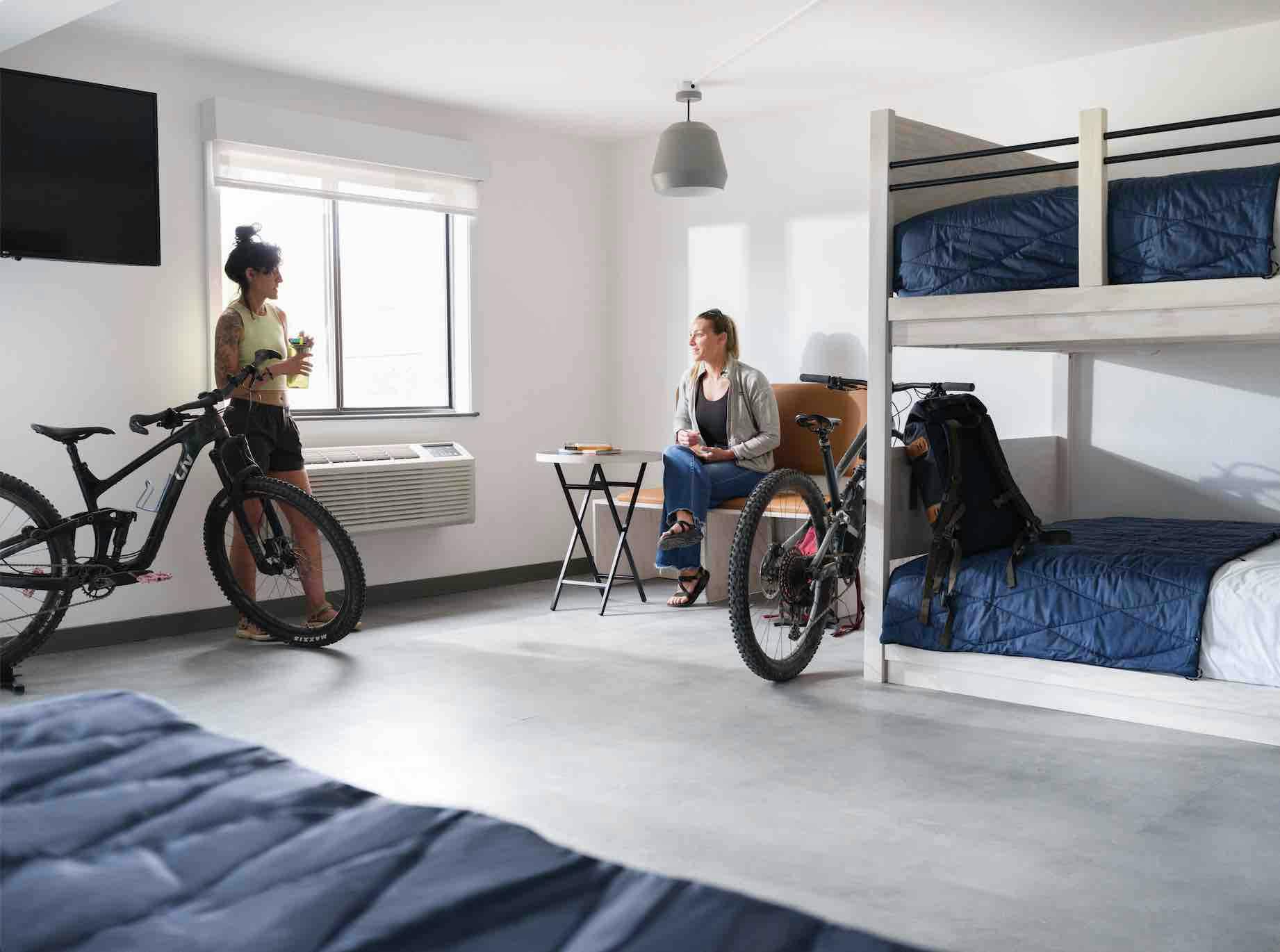 Two women are in a clean white hotel room with bunk beds. The women are having a conversation by the window, and there are 2 bikes in the room.